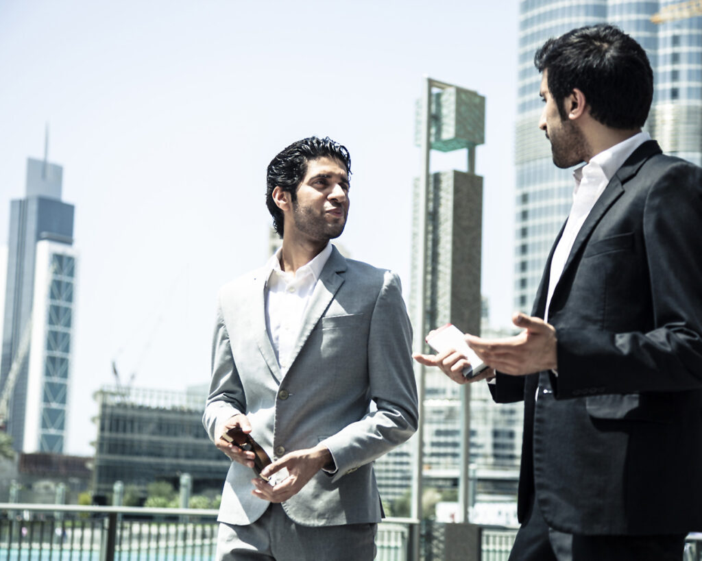 South Asian professional man standing against skyscrapers talking to another man in a suit.