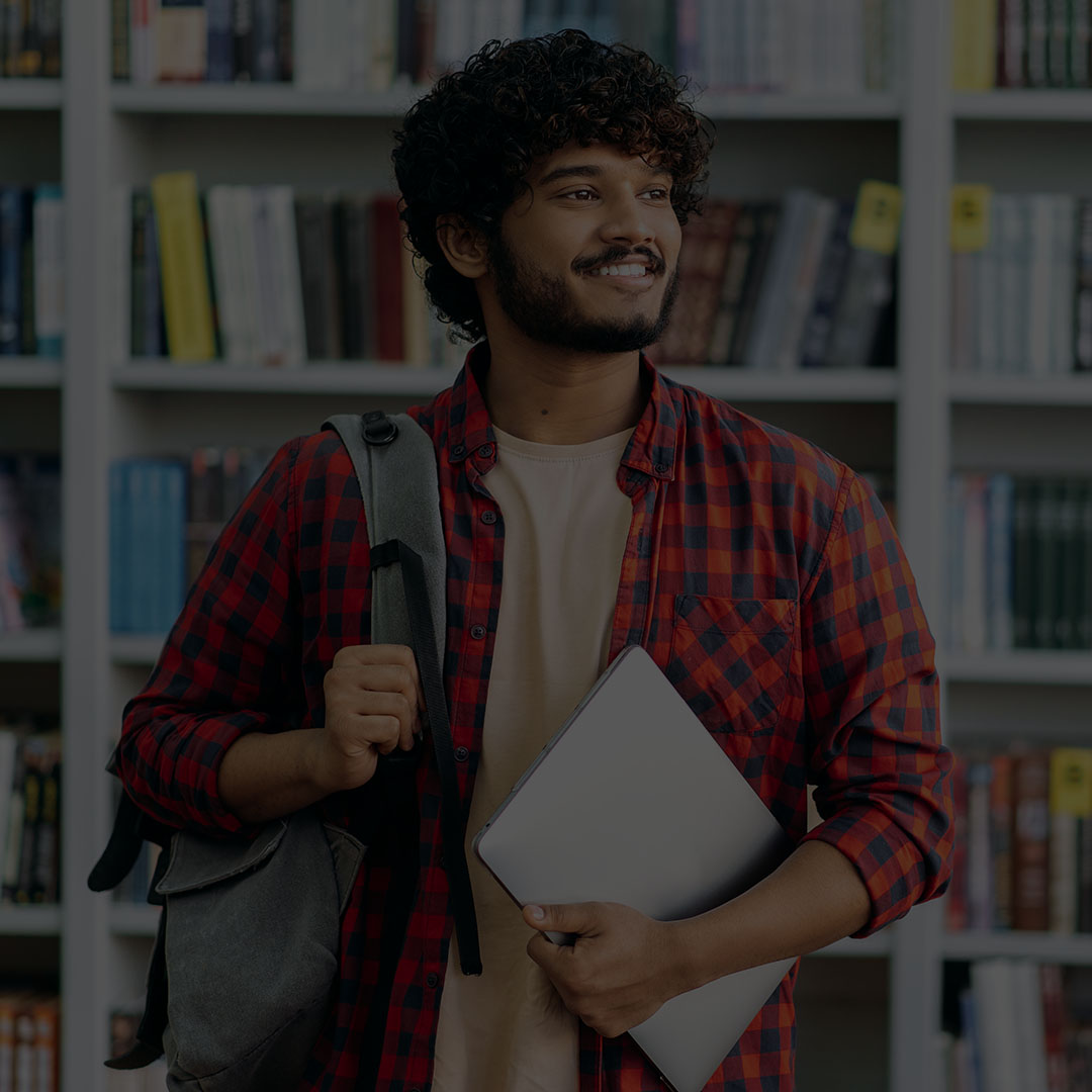 South Asian male student carrying a backpack and a book under his arm.