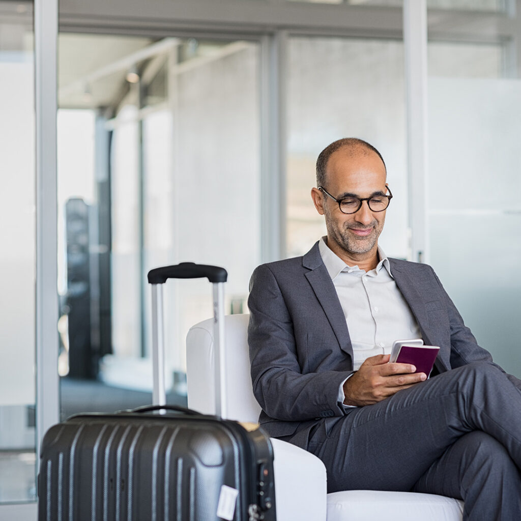 Man sitting at an airport looking at his phone with his suitcase next to him.