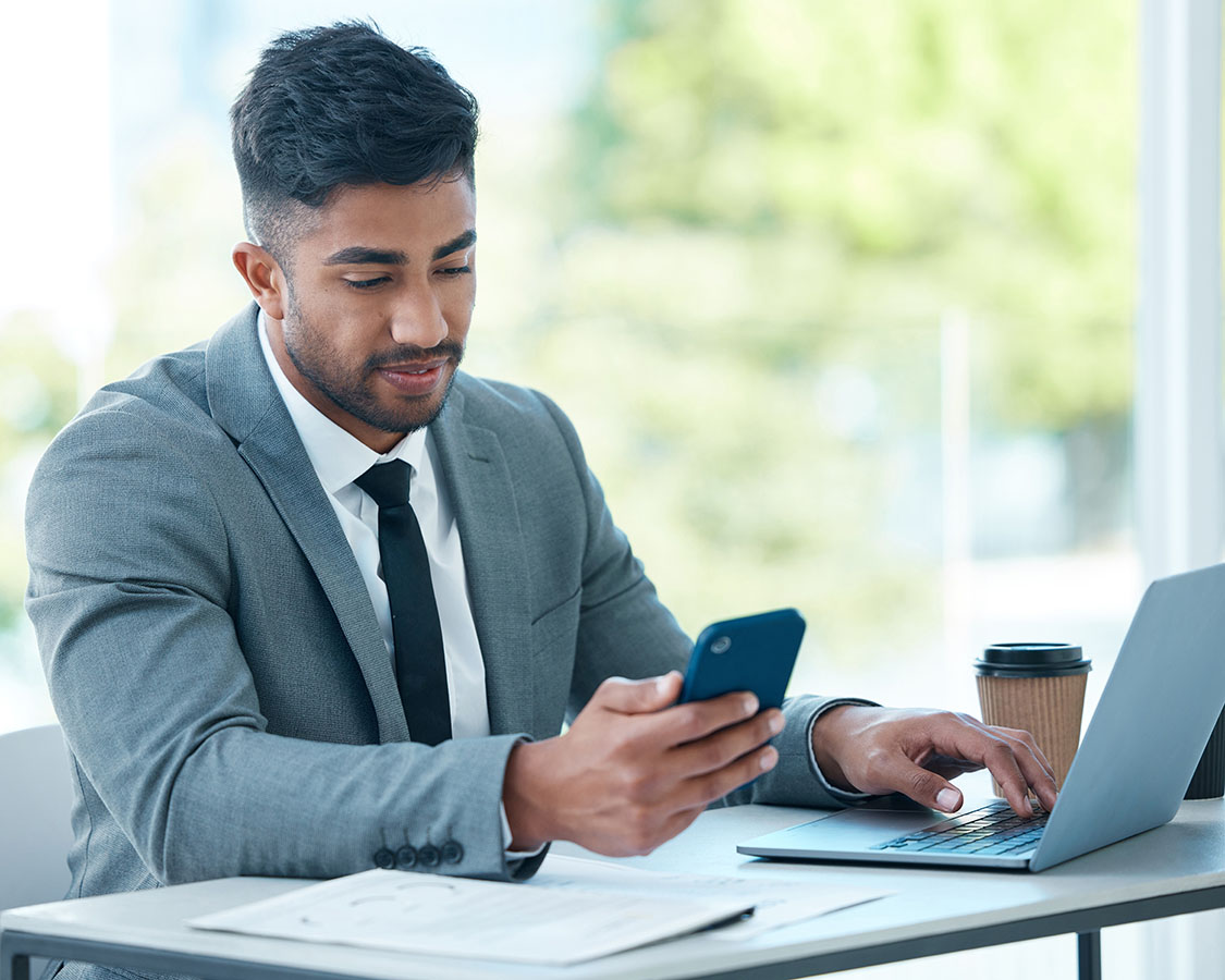 young business man at his desk with a laptop and coffee cup on the side, looking into his mobile phone.