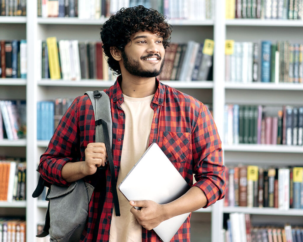 South Asian male student carrying a backpack and a book under his arm.