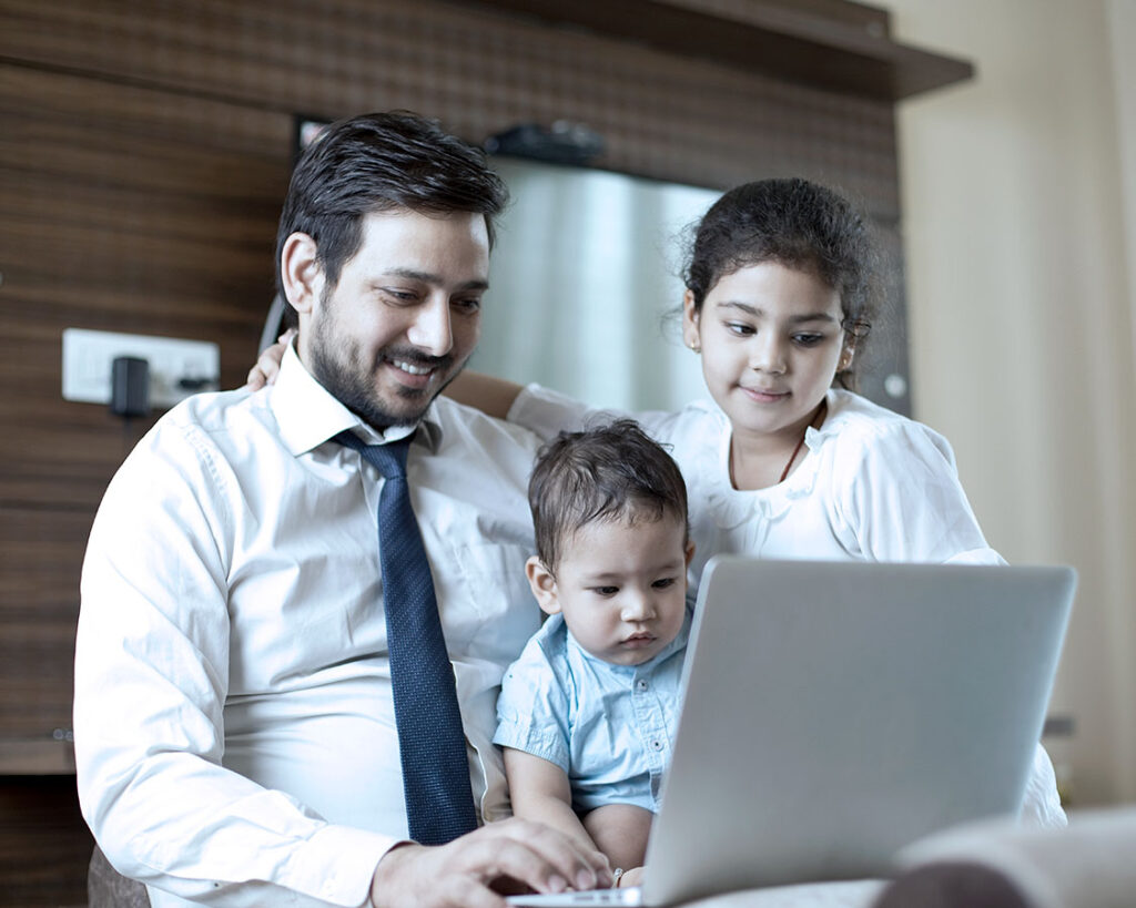 Man in a tie and button down shirt with 2 kids on his lap while working on his laptop.