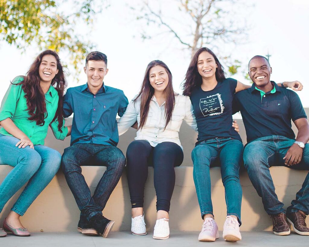 Multi ethnic students, male and female sitting on a concrete ledge smiling together.