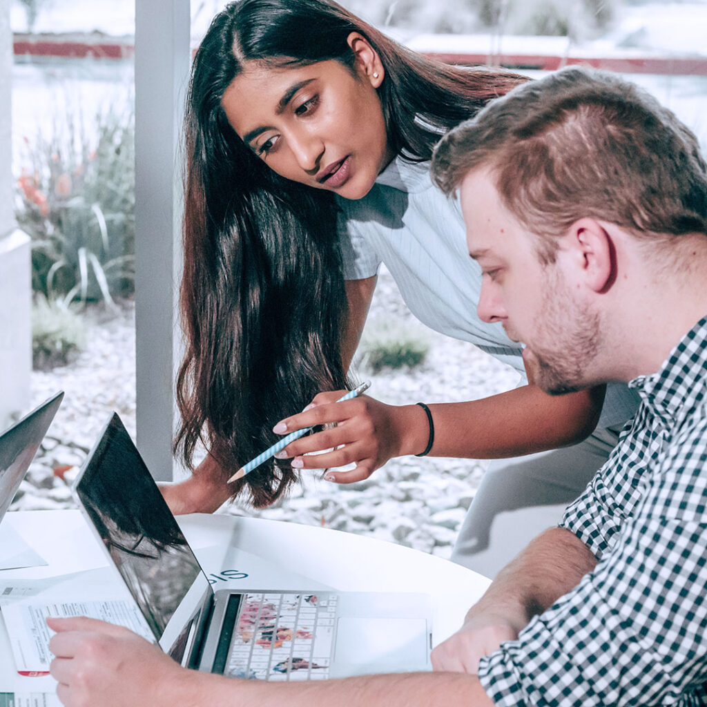 South Asian female leaning over a laptop with her fingers pointing to the screen with a caucasian male peering over the computer next to her.