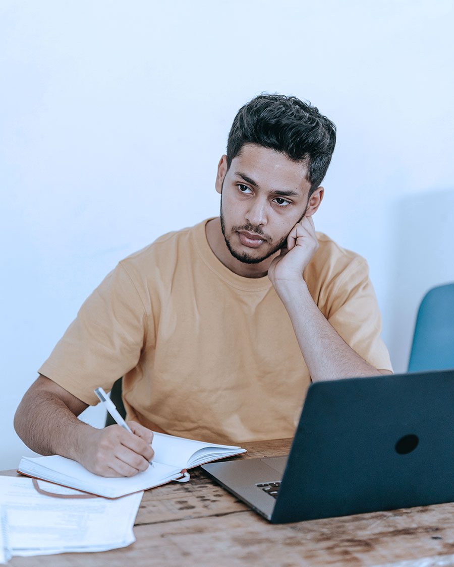 Ethnic student sitting at a desk in front of a laptop with pen in hand over a paper, looking away